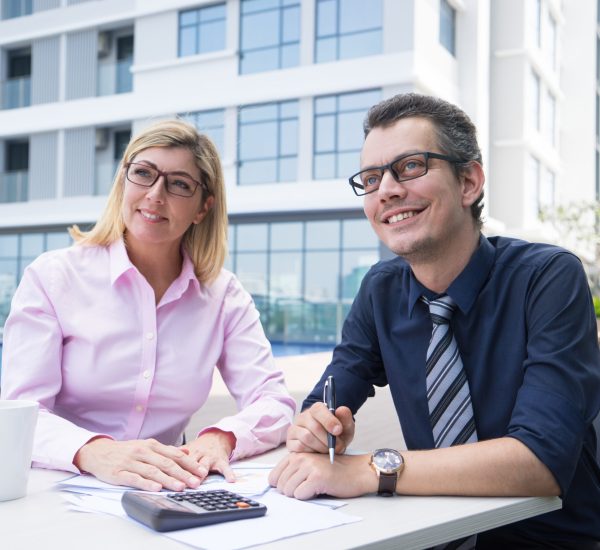 Smiling ambitious accountants contemplating while sitting at table with papers and calculator outdoors. Cheerful purposeful business colleagues in eyeglasses in formalwear working on budget planning. Financier concept